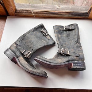 May include: A pair of distressed black leather boots with buckle closures. The boots have a worn, textured appearance with a gold-toned overlay. The soles are a dark red color, and the boots are set against a neutral background.