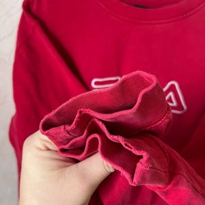 May include: A red sweatshirt with the white FILA logo on the front. The cuff of the sleeve is visible, showing the ribbed fabric. The sweatshirt is made of a soft material, perfect for casual wear.