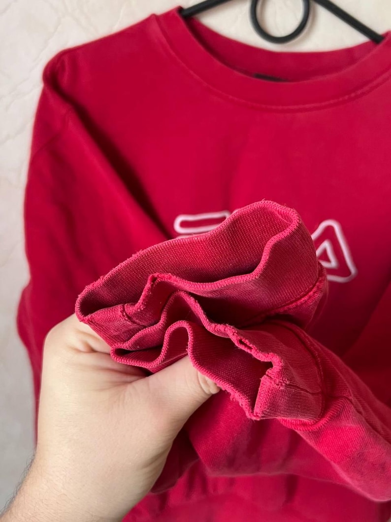 May include: A red sweatshirt with the white FILA logo on the front. The cuff of the sleeve is visible, showing the ribbed fabric. The sweatshirt is made of a soft material, perfect for casual wear.