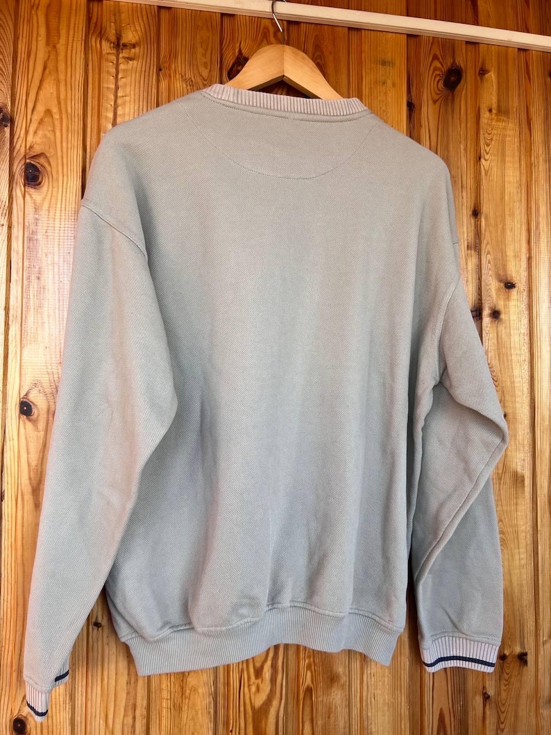 Puede incluir: Una sudadera verde claro con cuello redondo y pu&ntilde;os acanalados. Los pu&ntilde;os tienen un dise&ntilde;o a rayas con detalles en azul marino. La sudadera est&aacute; colgada en una percha de madera sobre un fondo de madera.