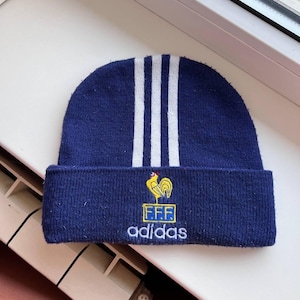 May include: A navy blue knitted beanie hat with three white stripes and a folded brim. The hat has a yellow rooster emblem with the letters "FFF" and the Adidas logo embroidered on the front. A winter accessory.