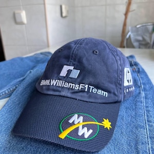May include: A navy blue baseball cap featuring the BMW WilliamsF1 Team logo embroidered in white on the front. The brim is made of a dark blue mesh material with a circular logo containing the letters MW and a yellow star graphic.