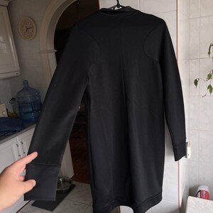 May include: A long-sleeved black dress with a high neckline and a fitted silhouette. The dress features a subtle seam detail on the shoulders and cuffs. The garment is hanging on a black hanger.