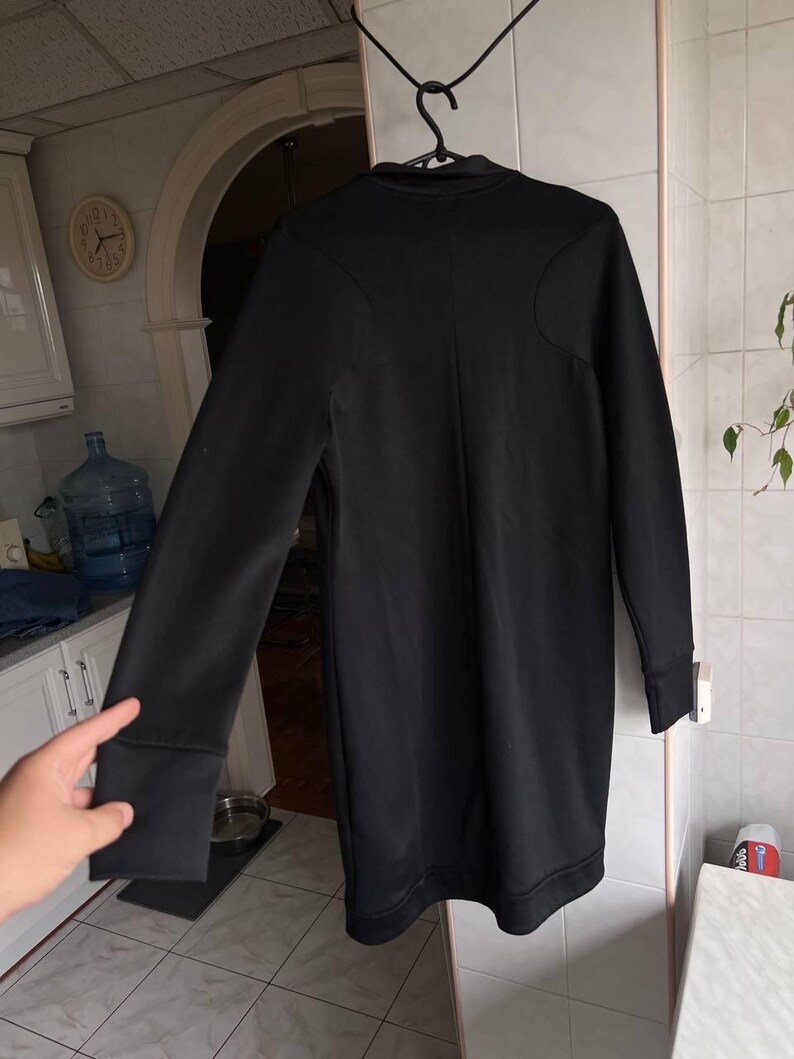 May include: A long-sleeved black dress with a high neckline and a fitted silhouette. The dress features a subtle seam detail on the shoulders and cuffs. The garment is hanging on a black hanger.