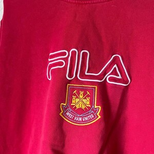 May include: A red FILA sweatshirt with a white outline logo and a West Ham United crest. The crest is a shield with crossed hammers and the words "West Ham United".