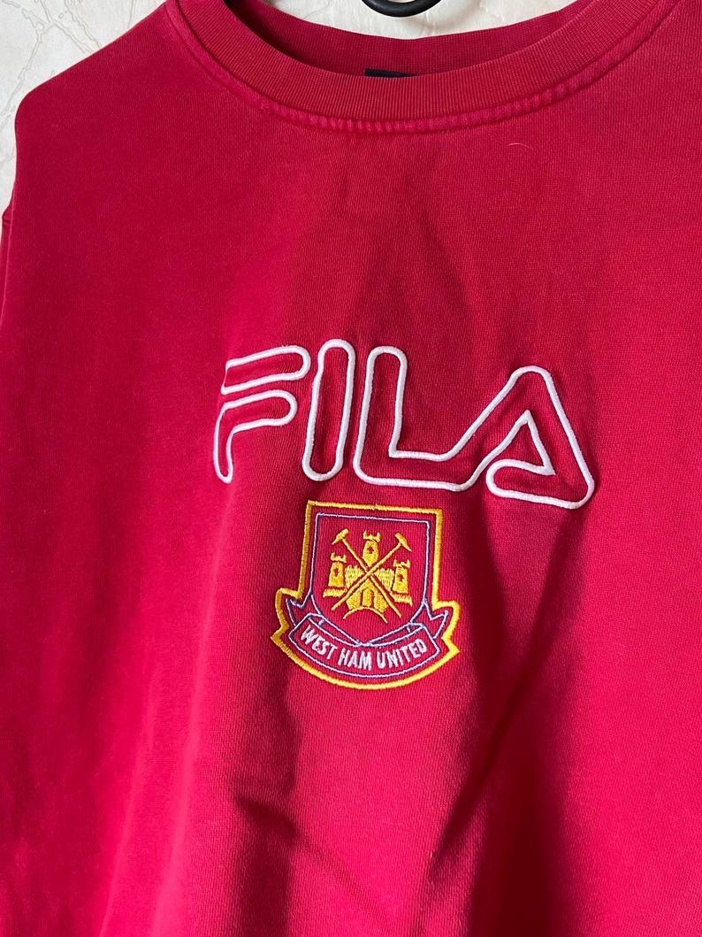May include: A red FILA sweatshirt with a white outline logo and a West Ham United crest. The crest is a shield with crossed hammers and the words "West Ham United".