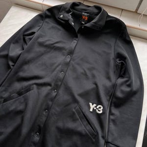 May include: Black button-up jacket with two front pockets and the white "Y-3" logo on the front. The jacket has a collared neckline and a small orange tag with the "Y-3" logo. The jacket is made of a smooth, shiny material.