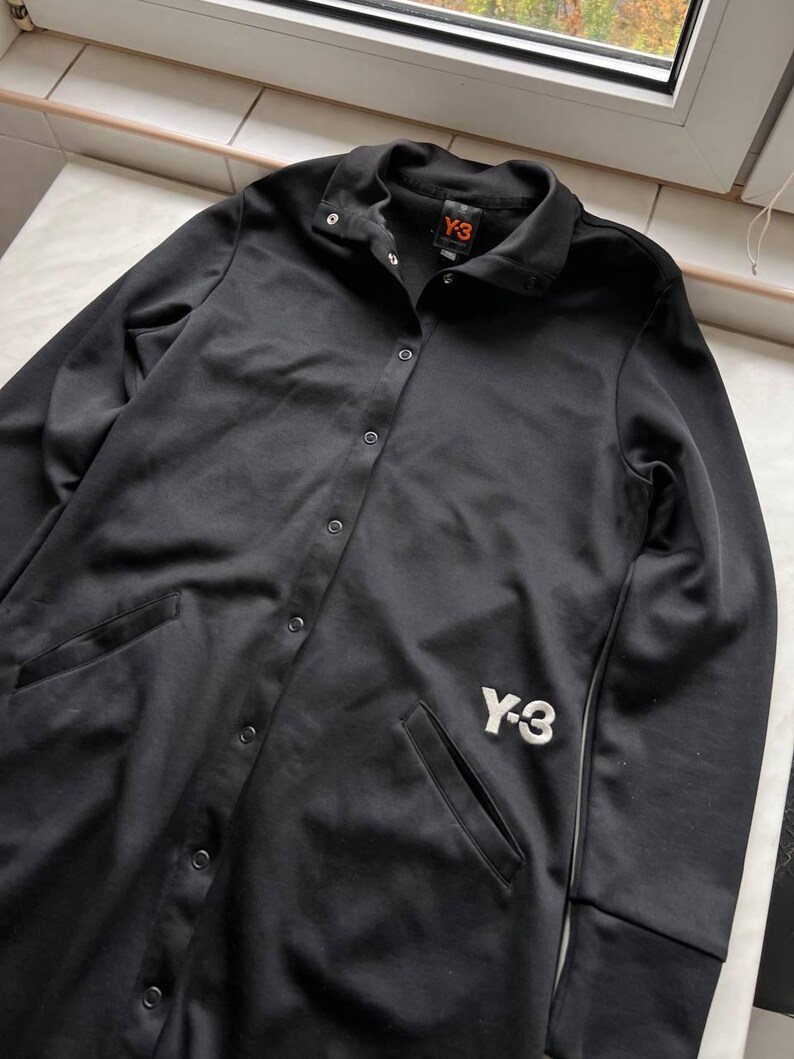 May include: Black button-up jacket with two front pockets and the white "Y-3" logo on the front. The jacket has a collared neckline and a small orange tag with the "Y-3" logo. The jacket is made of a smooth, shiny material.