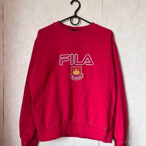 May include: A red FILA sweatshirt with a crew neck and long sleeves. The FILA logo is in white, outlined in red, and is centered on the chest. Below the logo is a crest with the words "WEST HAM UNITED".