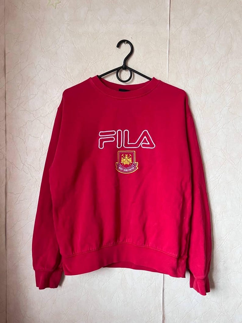 May include: A red FILA sweatshirt with a crew neck and long sleeves. The FILA logo is in white, outlined in red, and is centered on the chest. Below the logo is a crest with the words "WEST HAM UNITED".
