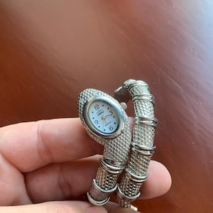 May include: A silver-toned snake-shaped watch with a textured, scale-like finish. The watch face is oval-shaped with a white background, black numerals at 12, 3, 6, and 9, and the text "USS" and "QUARTZ" printed in black on the dial.