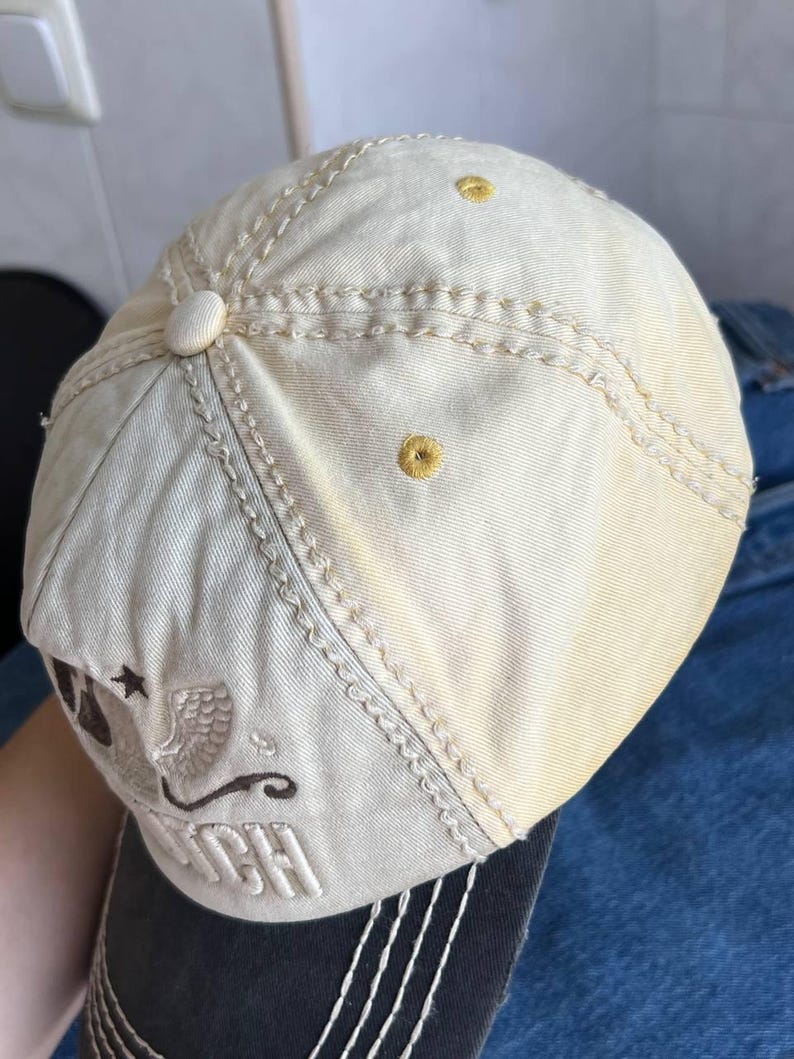 Op de afbeelding: Cr&egrave;mekleurige baseballpet met geborduurde details. Voorzien van een gebogen klep en decoratieve stiksels. De pet heeft een logo met het woord "Dutch" en een ster- en adelaarsontwerp.