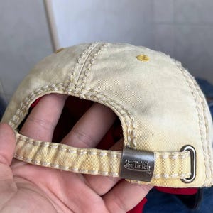 Op de afbeelding: Beige baseballpet met gele stiksels en een metalen sluiting. De pet heeft het "Von Dutch" logo op de sluiting. De binnenvoering is rood. De pet is gemaakt van een duurzame stof.