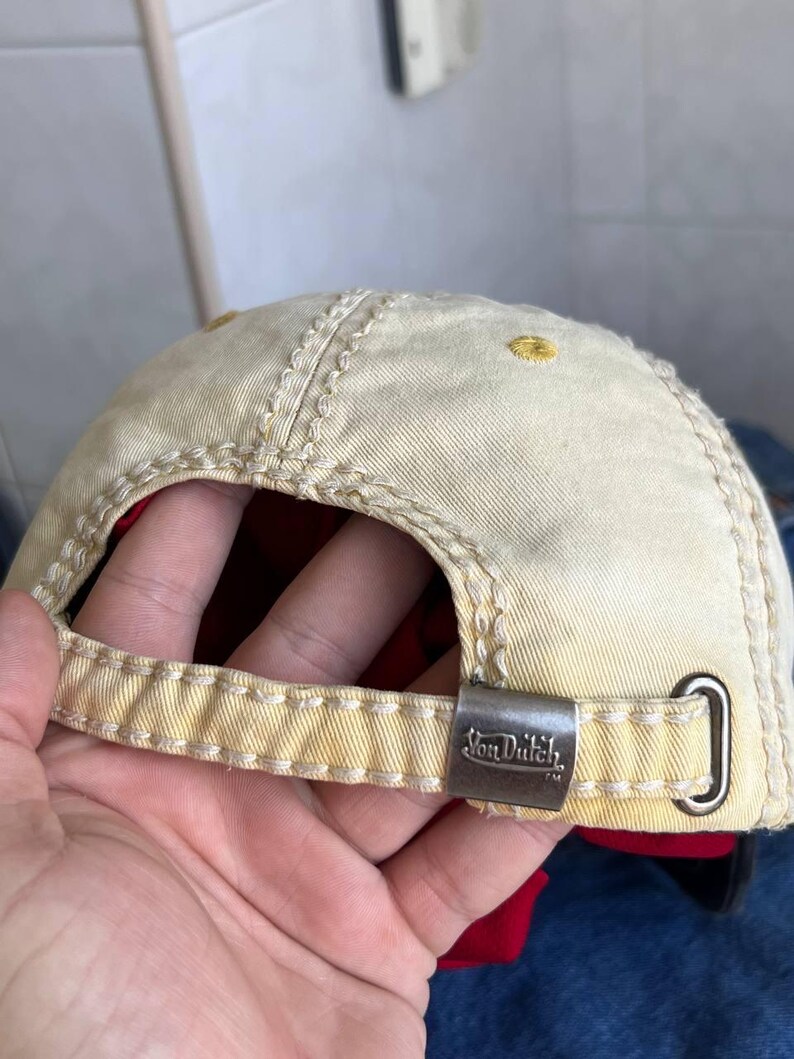 Op de afbeelding: Beige baseballpet met gele stiksels en een metalen sluiting. De pet heeft het "Von Dutch" logo op de sluiting. De binnenvoering is rood. De pet is gemaakt van een duurzame stof.