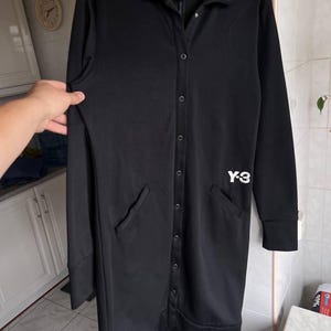 May include: Black long-sleeve button-up coat with two front pockets. The coat has a collar and the white "Y-3" logo on the chest. The coat is hanging on a black hanger.