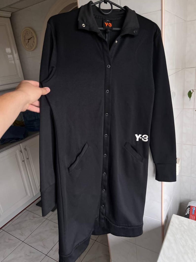 May include: Black long-sleeve button-up coat with two front pockets. The coat has a collar and the white "Y-3" logo on the chest. The coat is hanging on a black hanger.