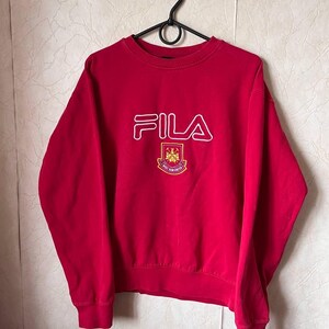 May include: A red FILA sweatshirt with a white outline logo and a crest. The crest features a maroon and gold emblem with the text "WEST HAM UNITED". The sweatshirt is hanging on a black hanger.