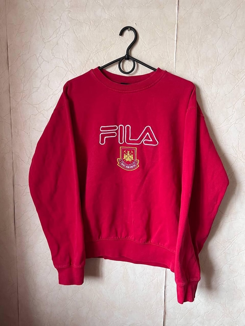 May include: A red FILA sweatshirt with a white outline logo and a crest. The crest features a maroon and gold emblem with the text "WEST HAM UNITED". The sweatshirt is hanging on a black hanger.