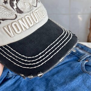 Op de afbeelding: Een distressed baseballcap met een zwarte klep en witte stiksels. De cap is beige met een geborduurd ontwerp en het woord "VONDUTCH" in het wit. De klep heeft een versleten, gerafelde rand.