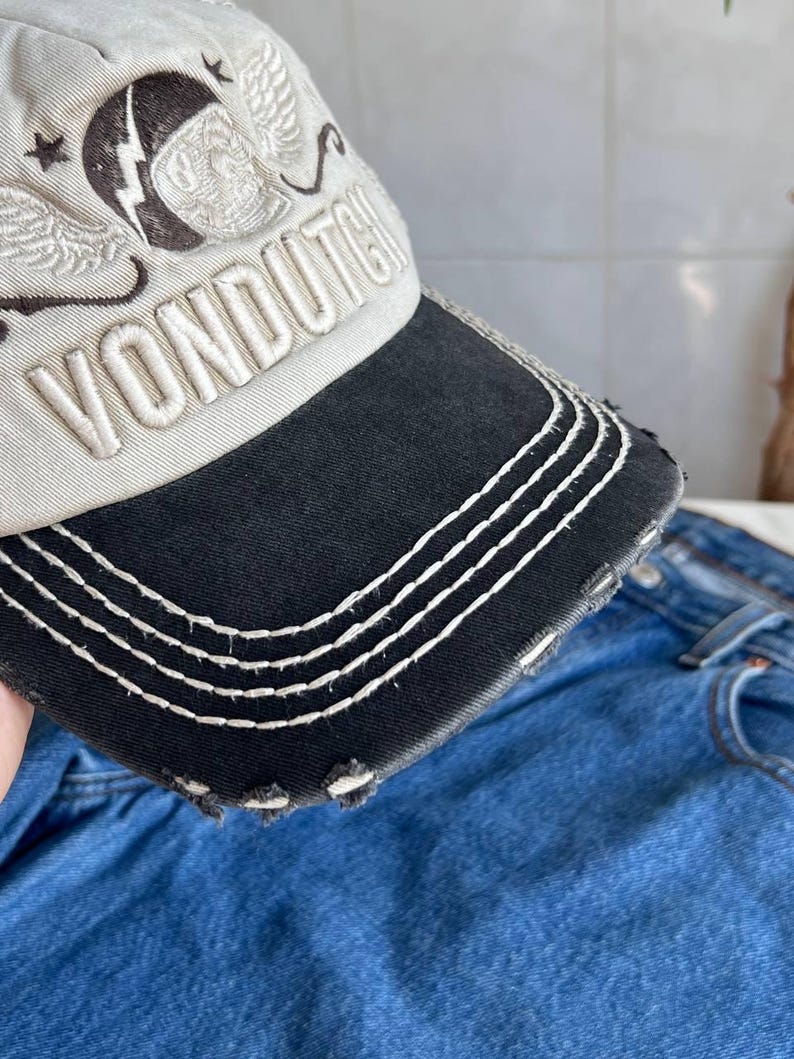 Op de afbeelding: Een distressed baseballcap met een zwarte klep en witte stiksels. De cap is beige met een geborduurd ontwerp en het woord "VONDUTCH" in het wit. De klep heeft een versleten, gerafelde rand.