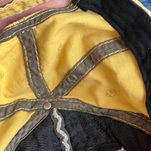 Op de afbeelding: Gele baseballpet met een zwarte rand en binnenkant. De pet heeft een grijze binnenvoering met de tekst "Von Dutch California" en een zilveren knoop. De binnenkant van de pet is geel.