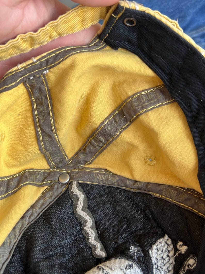 Op de afbeelding: Gele baseballpet met een zwarte rand en binnenkant. De pet heeft een grijze binnenvoering met de tekst "Von Dutch California" en een zilveren knoop. De binnenkant van de pet is geel.