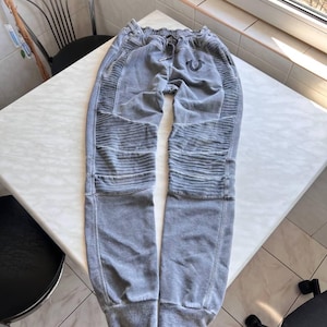 May include: Gray sweatpants featuring a faded wash, ribbed paneling on the thighs, and a drawstring waistband. The pants have a tapered leg design with cuffed ankles and a small black horseshoe logo embroidered near the upper left pocket area.