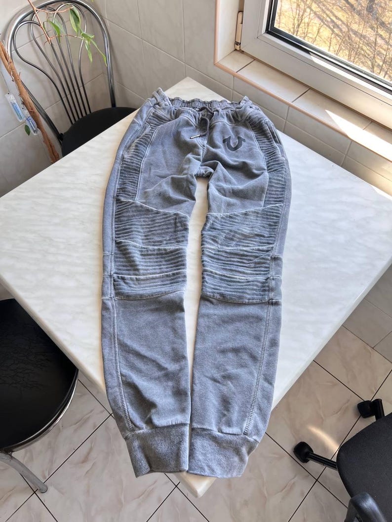May include: Gray sweatpants featuring a faded wash, ribbed paneling on the thighs, and a drawstring waistband. The pants have a tapered leg design with cuffed ankles and a small black horseshoe logo embroidered near the upper left pocket area.
