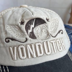 Op de afbeelding: Een beige baseballpet met een zwarte klep. De pet heeft de geborduurde tekst "VON DUTCH" en een bruin embleem met vleugels, sterren en een bliksemschicht. De pet heeft een gebogen klep en een knoop aan de bovenkant.