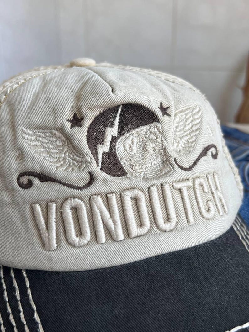 Op de afbeelding: Een beige baseballpet met een zwarte klep. De pet heeft de geborduurde tekst "VON DUTCH" en een bruin embleem met vleugels, sterren en een bliksemschicht. De pet heeft een gebogen klep en een knoop aan de bovenkant.