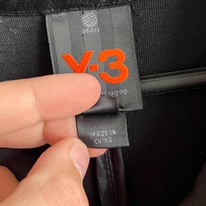 May include: Close-up of a black garment tag with the Adidas logo and the orange "Y-3" logo. The tag also includes the text "MADE IN CHINA". The garment appears to be made of a textured fabric.