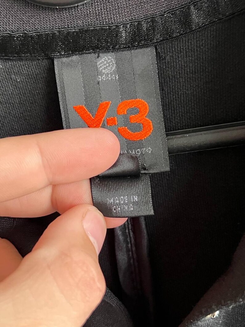 May include: Close-up of a black garment tag with the Adidas logo and the orange "Y-3" logo. The tag also includes the text "MADE IN CHINA". The garment appears to be made of a textured fabric.