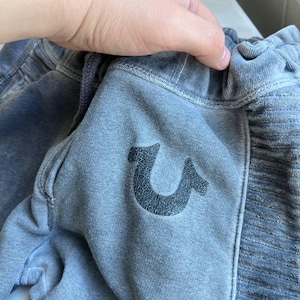 May include: Faded blue sweatpants featuring a dark, textured horseshoe logo on the front. The garment has an elasticized waistband with a drawstring and a ribbed side panel detail. The fabric has a soft, worn-in appearance with visible stitching.