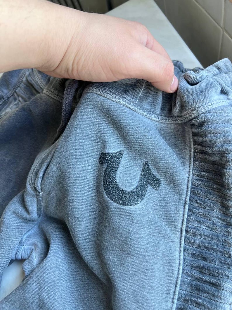 May include: Faded blue sweatpants featuring a dark, textured horseshoe logo on the front. The garment has an elasticized waistband with a drawstring and a ribbed side panel detail. The fabric has a soft, worn-in appearance with visible stitching.