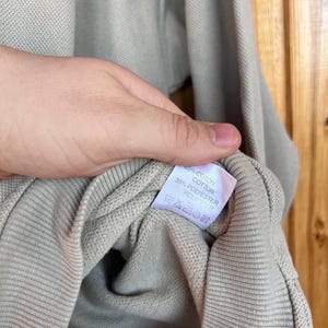 Puede incluir: Una camisa de manga larga de color beige claro con pu&ntilde;o acanalado. La etiqueta muestra la composici&oacute;n de la tela: 65% algod&oacute;n y 35% poli&eacute;ster. La camisa est&aacute; sujeta por una mano.