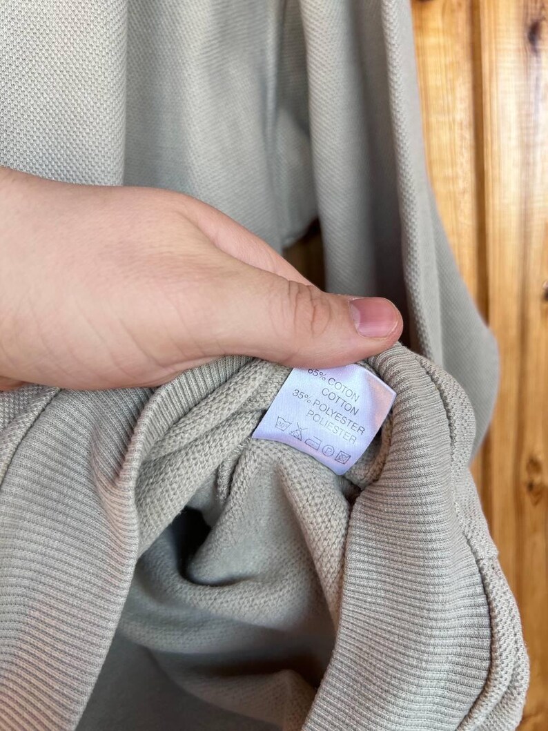Puede incluir: Una camisa de manga larga de color beige claro con pu&ntilde;o acanalado. La etiqueta muestra la composici&oacute;n de la tela: 65% algod&oacute;n y 35% poli&eacute;ster. La camisa est&aacute; sujeta por una mano.
