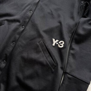May include: Black button-up jacket with a white embroidered "Y-3" logo on the front. The jacket has a pocket on the front and a ribbed cuff on the sleeve. The jacket is made of a smooth, matte fabric.