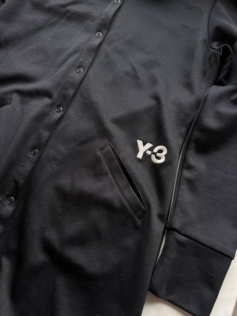May include: Black button-up jacket with a white embroidered "Y-3" logo on the front. The jacket has a pocket on the front and a ribbed cuff on the sleeve. The jacket is made of a smooth, matte fabric.