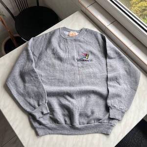 以下が含まれることがあります： 左胸にMicrosoft Windows 95の小さな刺繍ロゴが入った、グレーのヘザーカラーのクルーネックトレーナー。ロゴには、クラシックなマルチカラーのウィンドウアイコンと、黒い文字でWindows 95というテキストがデザインされています。