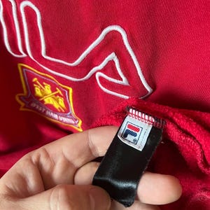 May include: Red sweatshirt with a white embroidered FILA logo. The garment features a team crest and a black tag with the FILA logo. The fabric has a textured appearance.