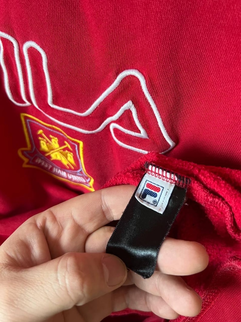May include: Red sweatshirt with a white embroidered FILA logo. The garment features a team crest and a black tag with the FILA logo. The fabric has a textured appearance.