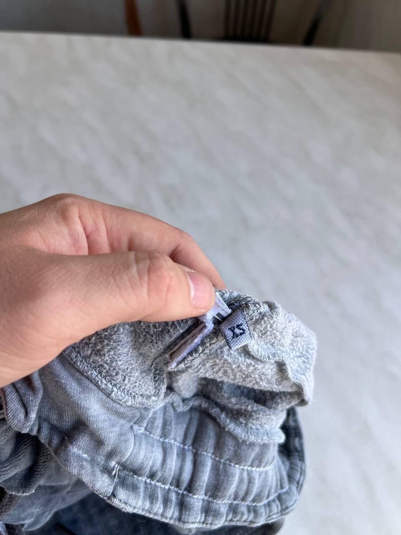 May include: A pair of grey sweatpants featuring an elasticized waistband with visible stitching. A small fabric tag is attached to the interior of the waistband, clearly displaying the size label XS in dark blue text on a light grey background.