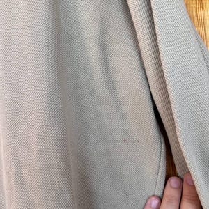 Puede incluir: Primer plano de un polo de color beige claro. La camisa tiene un tejido texturizado y una peque&ntilde;a mancha. La palabra "DOS" es visible en letras blancas en la parte superior. La manga y parte de la camisa son visibles.