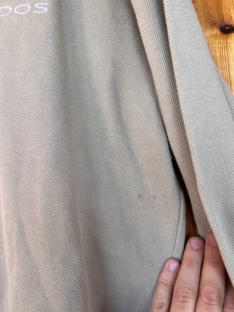 Puede incluir: Primer plano de un polo de color beige claro. La camisa tiene un tejido texturizado y una peque&ntilde;a mancha. La palabra "DOS" es visible en letras blancas en la parte superior. La manga y parte de la camisa son visibles.