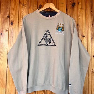 Puede incluir: Sudadera beige con cuello redondo y manga larga. La sudadera presenta un logotipo triangular negro con un gallo, un escudo del equipo y la palabra "EIDOS". Los pu&ntilde;os y el dobladillo tienen una raya azul marino y blanca.