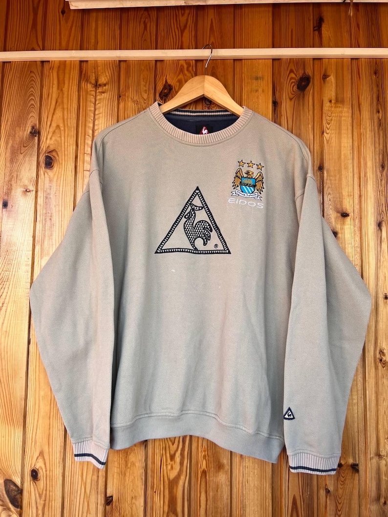 Puede incluir: Sudadera beige con cuello redondo y manga larga. La sudadera presenta un logotipo triangular negro con un gallo, un escudo del equipo y la palabra "EIDOS". Los pu&ntilde;os y el dobladillo tienen una raya azul marino y blanca.