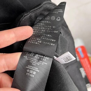 May include: Close-up of a black garment tag with Japanese and Chinese text. The tag includes care symbols and the text "MADE IN CHINA FABRIQUE EN CHINE". The tag is held by a hand, with a blurred background.