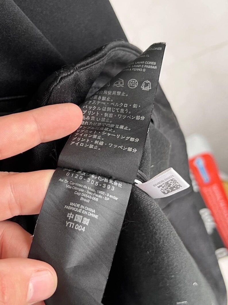May include: Close-up of a black garment tag with Japanese and Chinese text. The tag includes care symbols and the text "MADE IN CHINA FABRIQUE EN CHINE". The tag is held by a hand, with a blurred background.