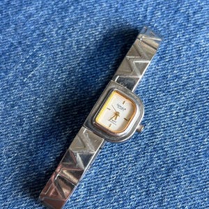 May include: A silver-tone Omax Quartz wristwatch featuring a rectangular face with a gold-coloured inner border and a white dial. The watch band is composed of metallic links, each embossed with a repeating geometric zigzag pattern.