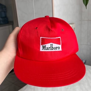 Puede incluir: Gorra de béisbol roja brillante con visera plana. La gorra presenta un parche rectangular con la palabra "Marlboro" en letras blancas. El parche tiene un diseño rojo y blanco.